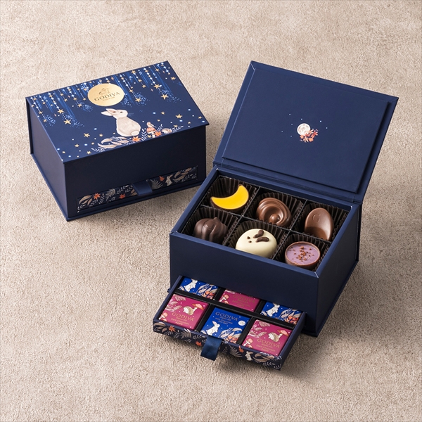 Opened navy-blue Godiva Autumn Collection box showing assorted chocolates in the main compartment and a lower drawer with individually wrapped chocolate squares.