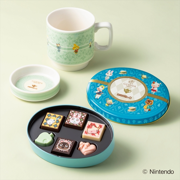An open blue tin of six assorted chocolates displayed alongside a pale green lidded tea mug and a small saucer on a light background.