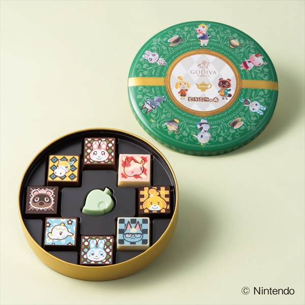 Opened green Godiva "Animal Crossing" assortment tin showing nine character-decorated chocolates in a tray with the tin lid beside it.