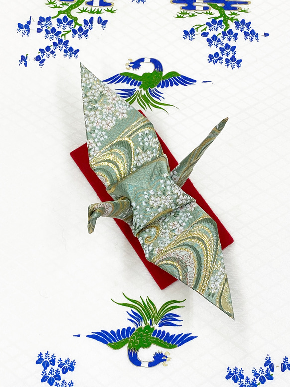 Top-down photo of the 折り鶴 figure in pale green cherry‑blossom and flowing‑water pattern placed on a red cushion.