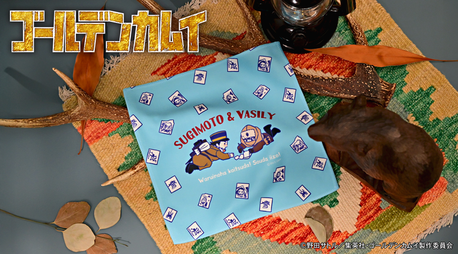 Light blue hand towel with chibi illustrations of Sugimoto Saichi and Vasily and the text "SUGIMOTO & VASILY" surrounded by small illustrated cards, laid on a woven mat.