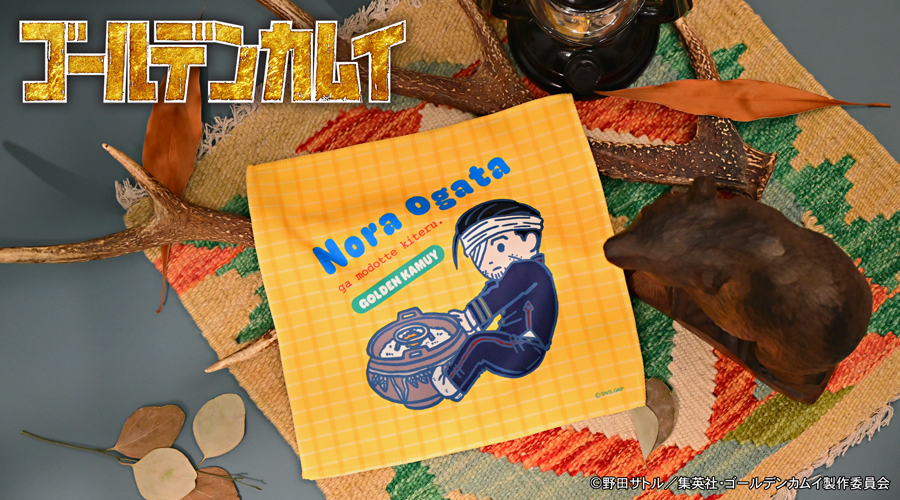 Folded yellow-check hand towel showing an illustration of Nora Ogata, placed on a tabletop with decorative items.