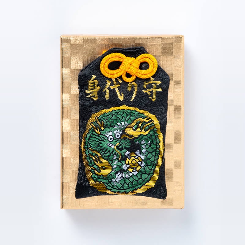 Embroidered black omamori amulet with gold embroidery and a yellow cord, displayed on a gold patterned box.