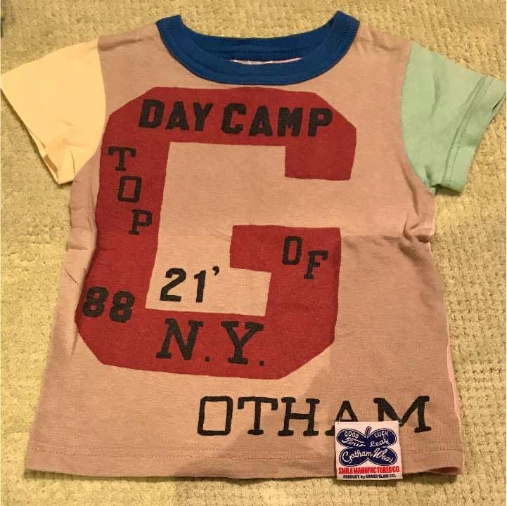 Children's cotton shirt laid flat showing the front with multicolored sleeves, large red 'G' graphic reading 'DAY CAMP', and a small brand patch at the hem.