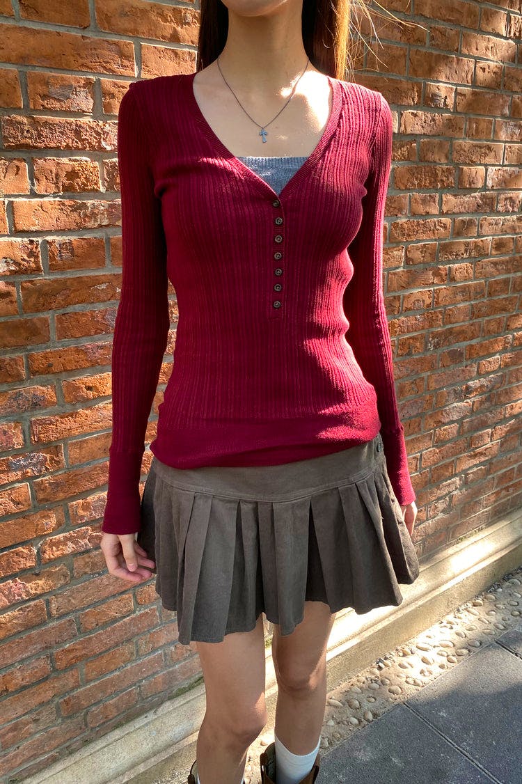 Front view of a red ribbed V-neck sweater with a buttoned placket worn by a model standing against a brick wall.