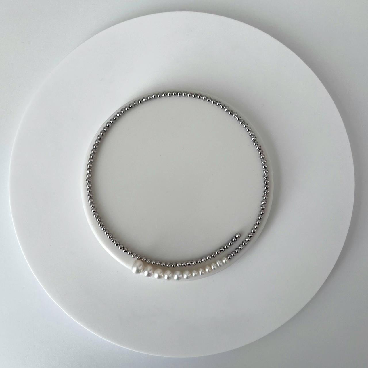 A silver gradation pearl choker laid flat on a white round display, showing small silver beads with a short row of graduated pearls.