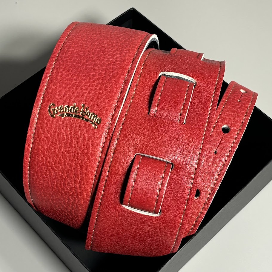 Textured red guitar strap with a gold "Grande Uomo" logo coiled on a black box.