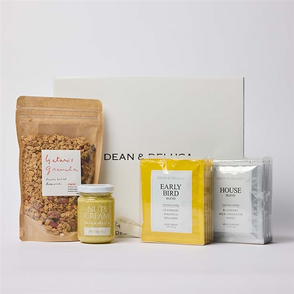 A granola and coffee gift set arranged in front of a DEAN & DELUCA box, showing a pouch of granola, a small jar of nut cream, and two packets of coffee.