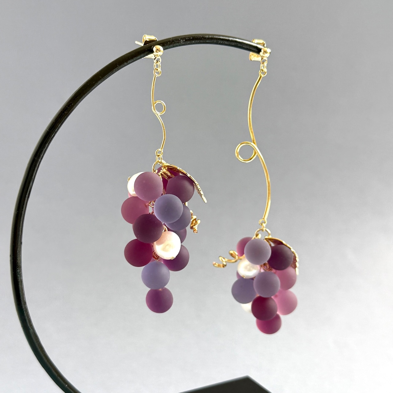 Grape-shaped drop earrings with clustered matte purple beads on gold-tone hooks shown on a jewelry stand.