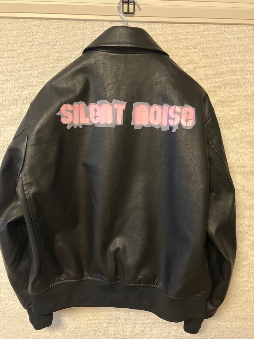 Back view of a men's synthetic leather zip-up blouson jacket with pink "SILENT NOISE" painted text on the back, hanging on a hook.