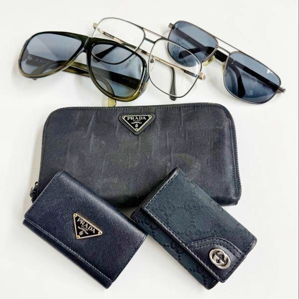 A set of branded accessories on a white background: a Prada zip-around wallet, two small leather key/card cases (one with a Gucci logo), and several sunglasses.