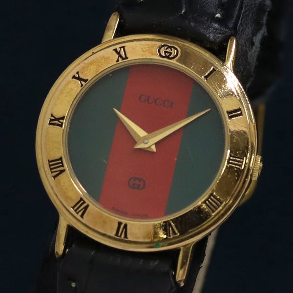 Front view of a Gucci women's wristwatch showing a green-and-red striped dial, gold bezel with Roman numerals, and a black leather strap.