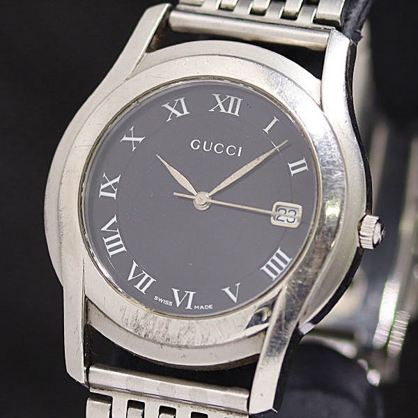 Close-up of the GUCCI 5500M men's wristwatch showing its black dial, Roman numerals, date window, and stainless-steel case and bracelet.