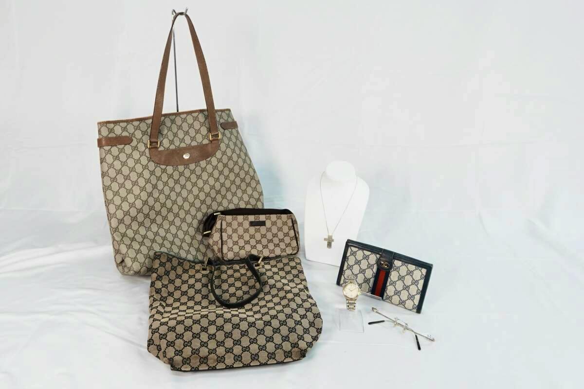 A GUCCI set arranged on white background including a monogram tote bag, two smaller monogram bags, a wallet, a wristwatch, eyeglasses, and a necklace on a display bust.