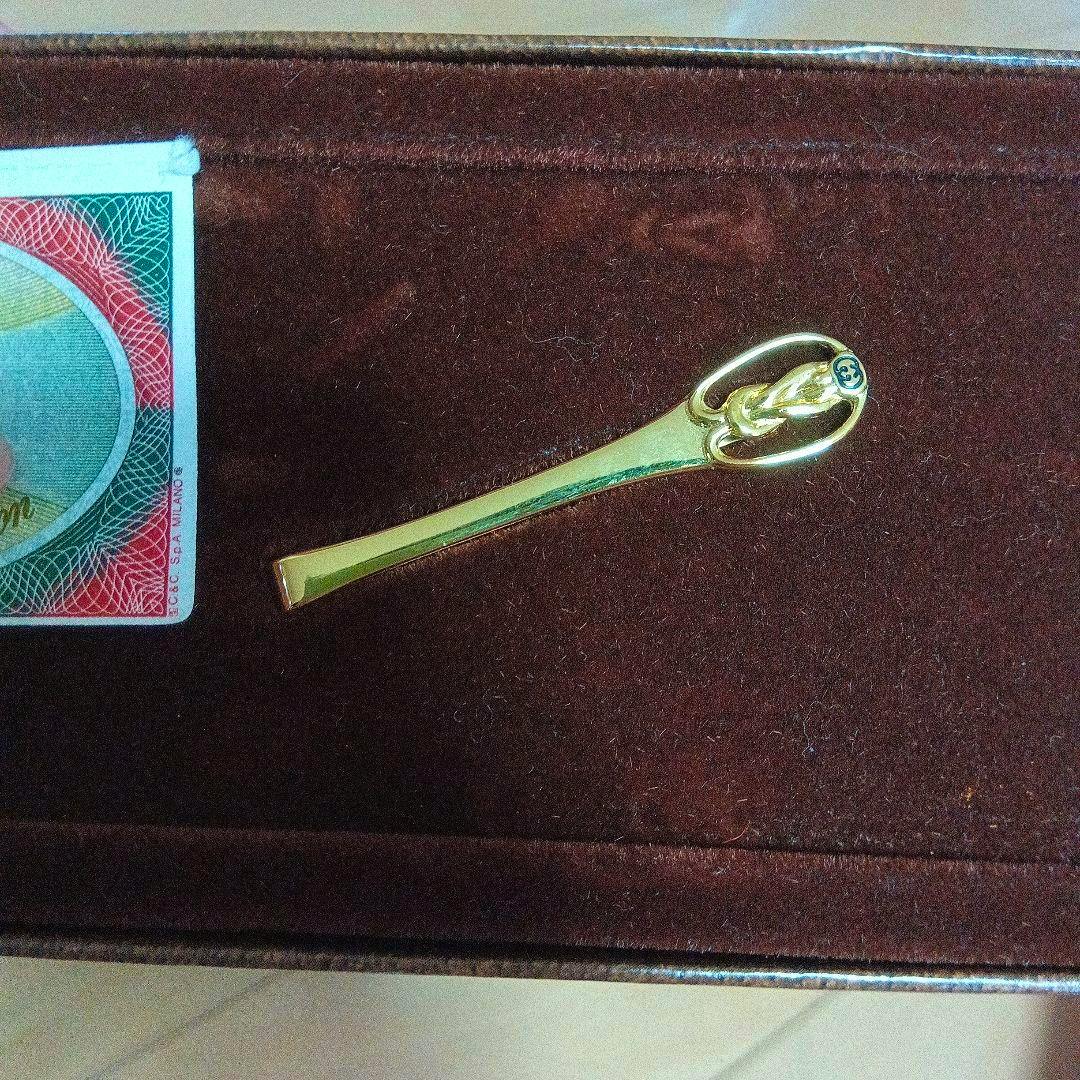 A gold-colored GUCCI tie pin displayed on brown velvet with a GUCCI accessory card visible.