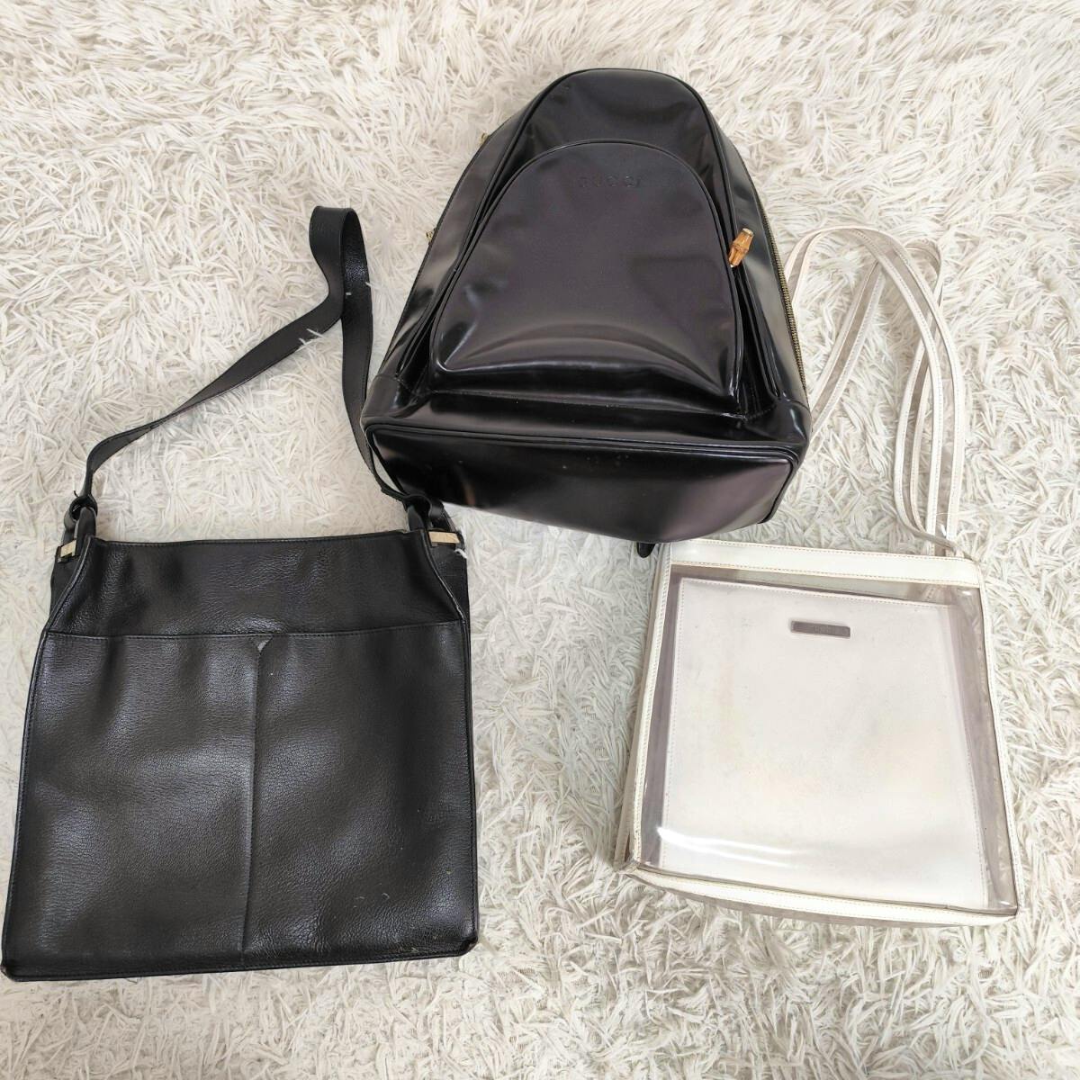 Three GUCCI bags laid on a light rug: a black shoulder bag, a black shiny backpack, and a white clear-structured shoulder bag.