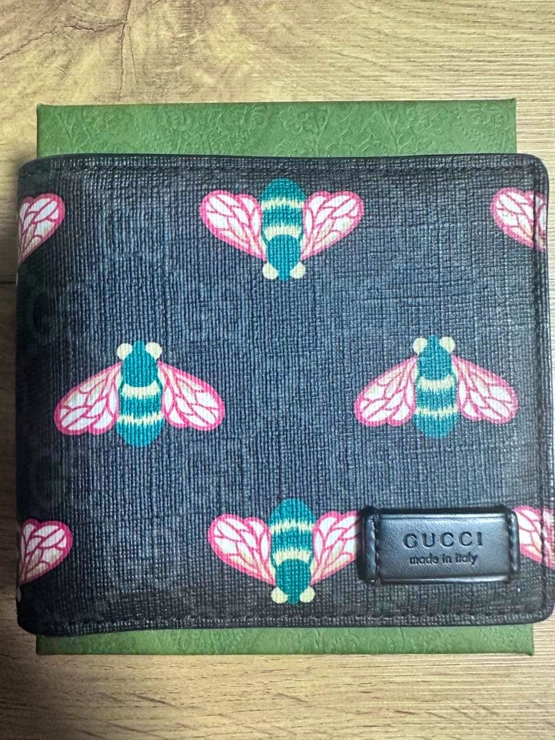 Canvas Gucci bi-fold wallet front with repeated pink-and-teal bee motif and a leather "GUCCI made in italy" patch.