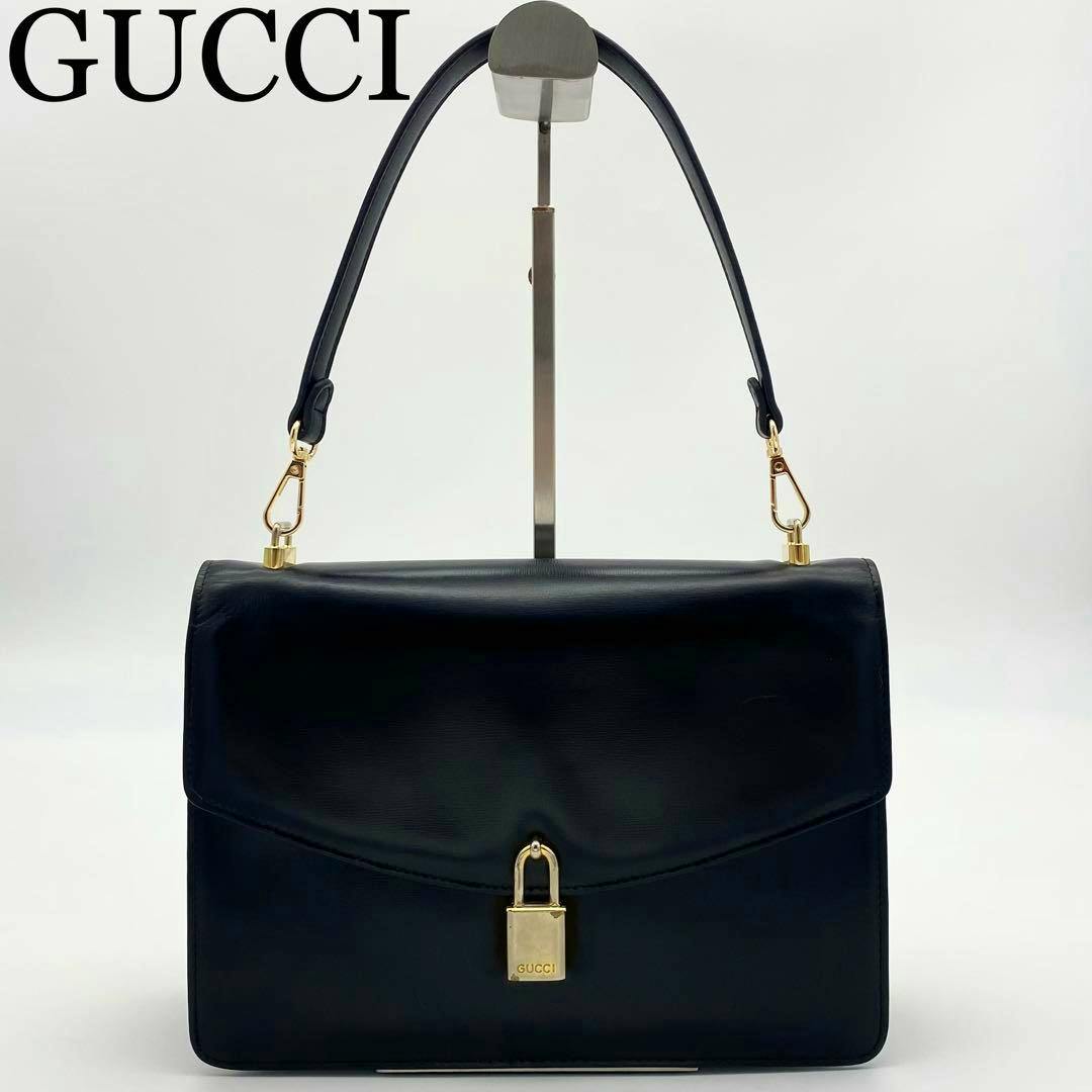 Front view of a black leather GUCCI handbag with gold padlock hardware and a top handle on a display stand.