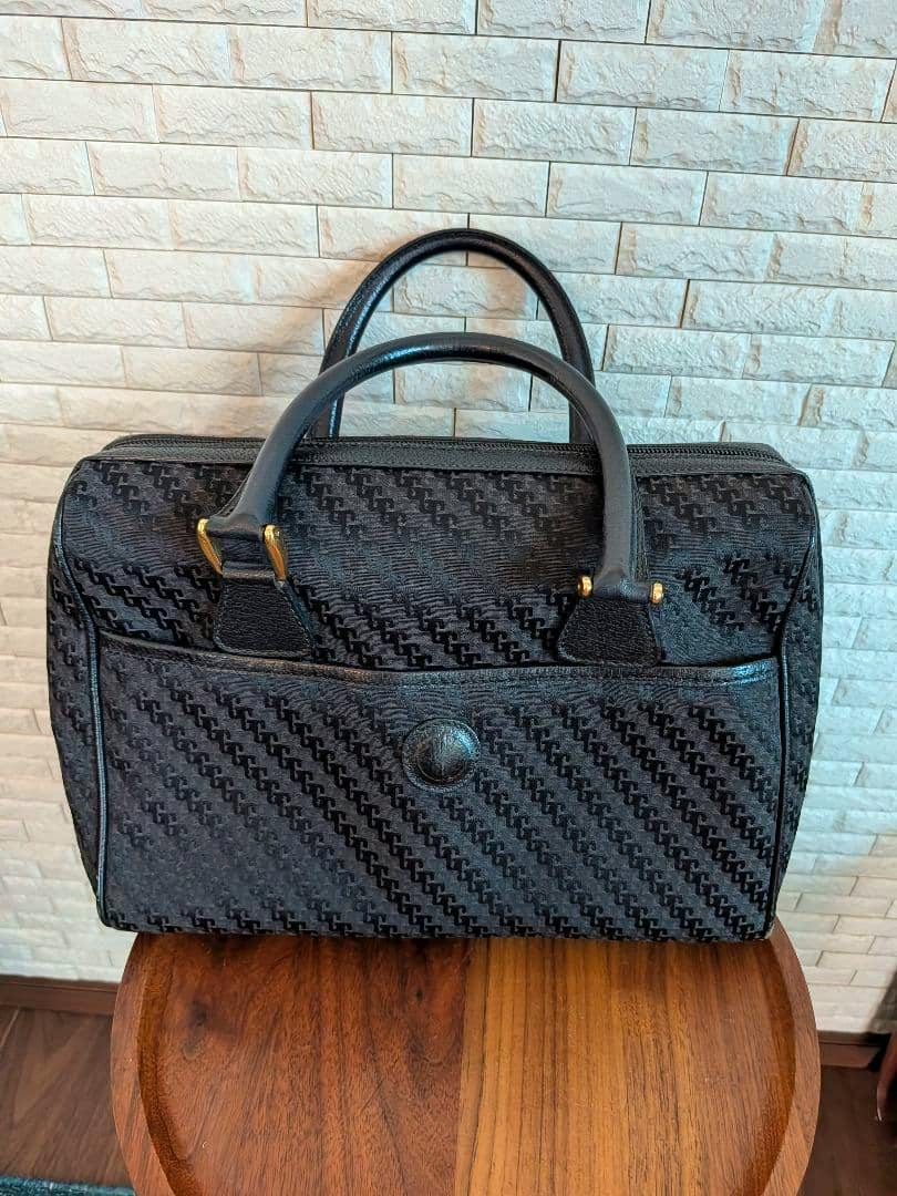 Black canvas Gucci handbag with all-over pattern, two top handles and a front pocket with a snap, photographed on a wooden stool against a tiled wall.