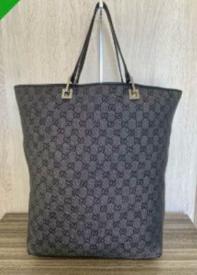 Front view of a black Gucci GG-pattern canvas tote bag with leather straps hanging against a wooden background.