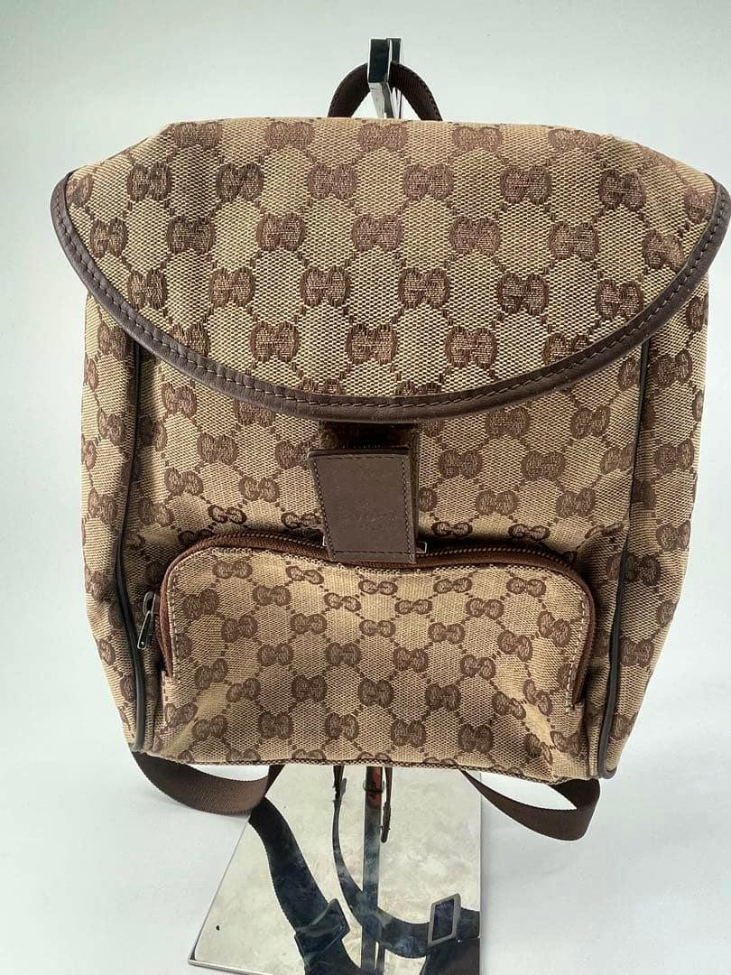 Front view of a GUCCI GG canvas backpack in beige and brown with a front zip pocket, displayed on a stand.