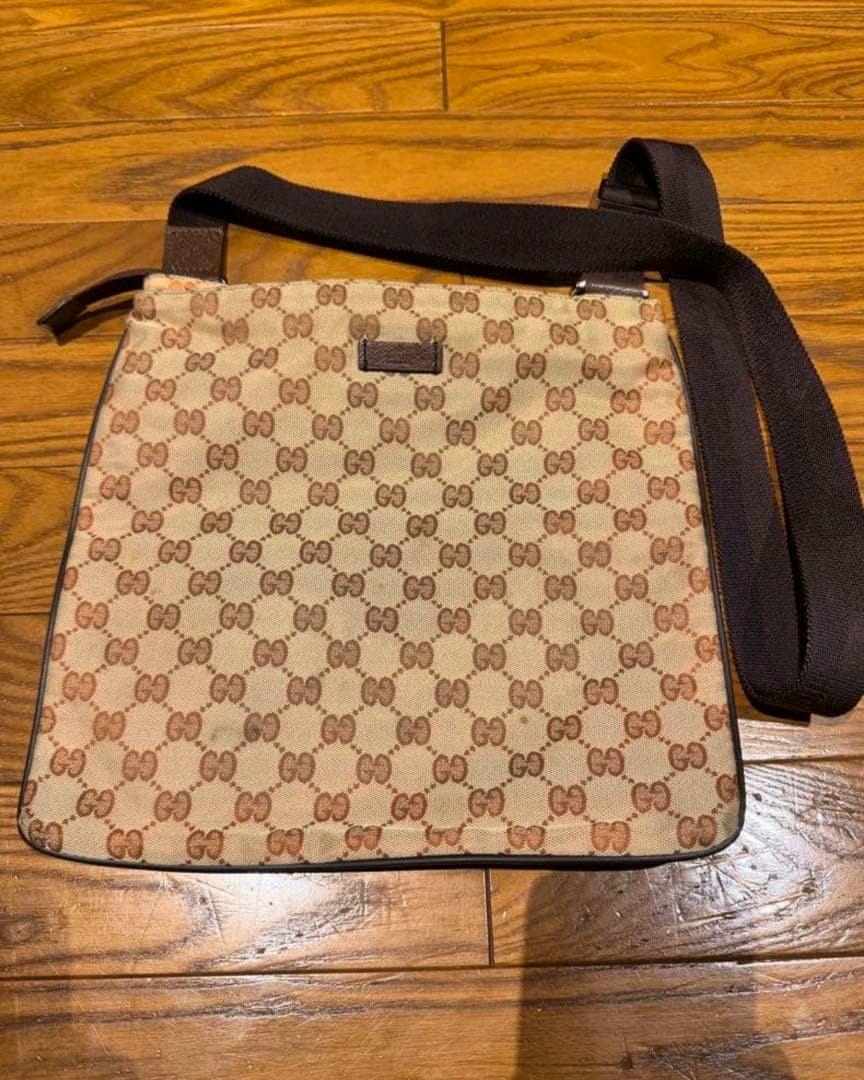 A brown Gucci GG canvas shoulder bag with a dark brown shoulder strap laid flat on a wooden floor.