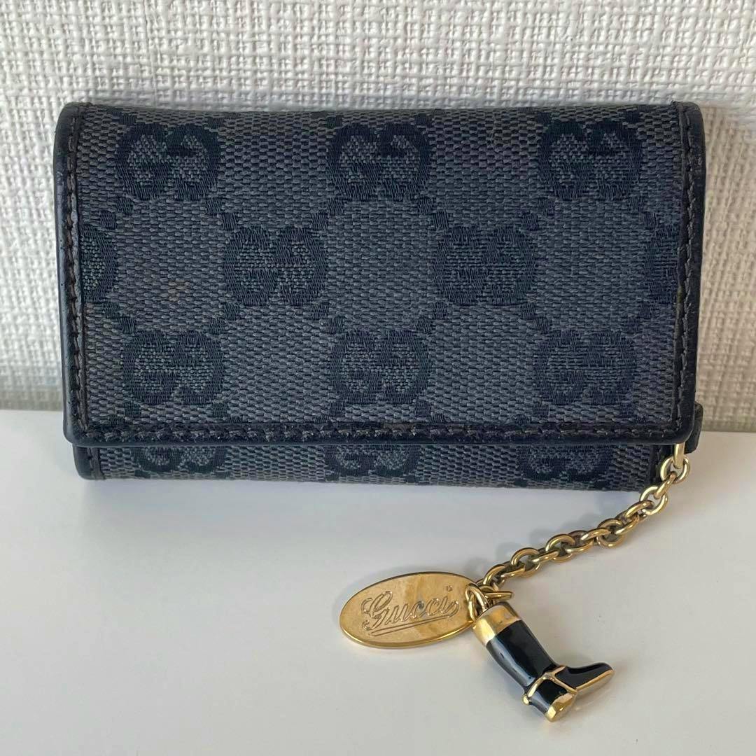 Black Gucci GG canvas key case shown front-facing with an attached gold-tone chain and boot-shaped charm plus an oval Gucci tag.