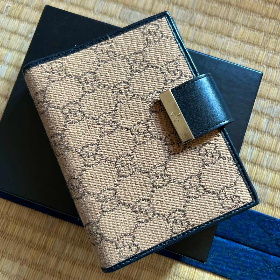 A Gucci GG canvas notebook cover with a black leather strap and metal clasp, shown resting on a black box.