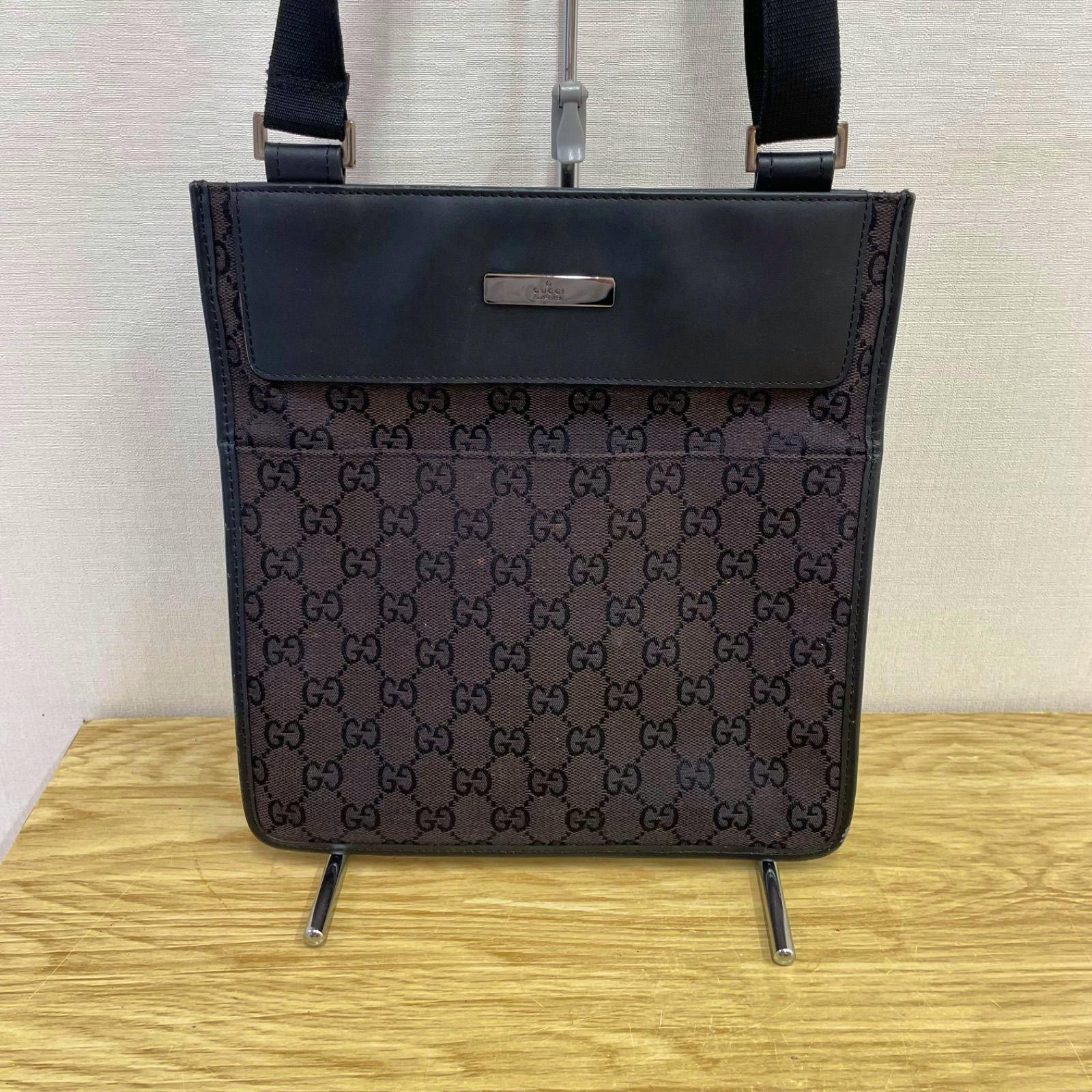 Front view of a brown Gucci GG canvas shoulder bag with a leather flap and metal logo plate, hanging on a stand.
