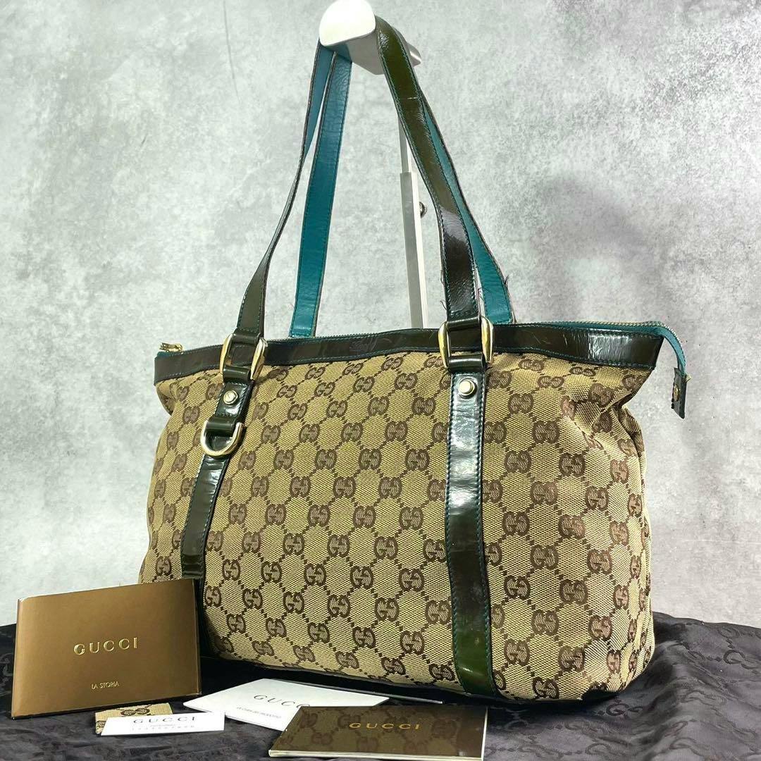 Brown Gucci GG canvas tote bag with dark leather trim and shoulder straps displayed with Gucci booklets in front.
