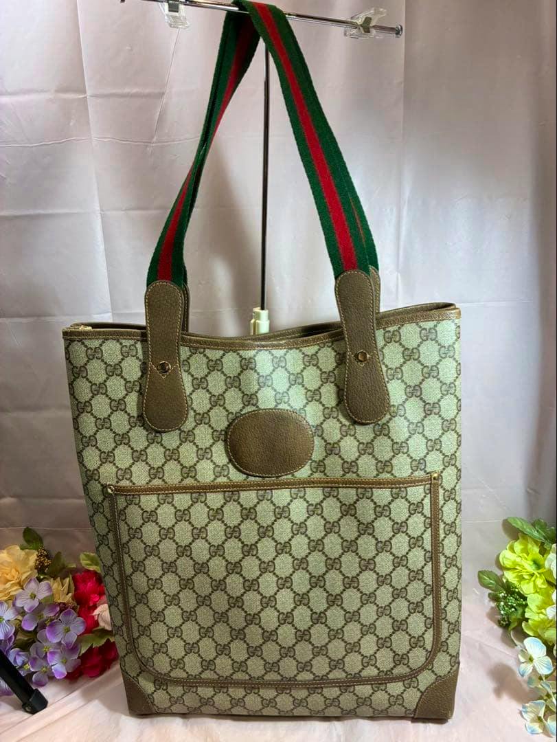 Beige Gucci GG monogram canvas tote bag with green-red-green shoulder straps and brown leather trim shown upright on a stand.