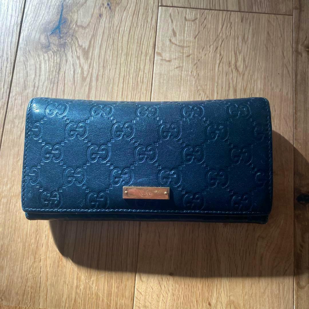 Closed black Gucci long leather wallet with embossed GG pattern and a small rectangular metal plate on a wooden surface.