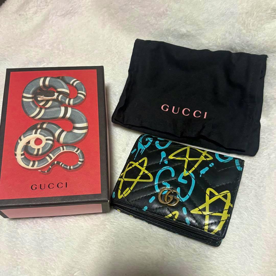 A Gucci card case wallet with blue and yellow graffiti-style print and a gold GG logo shown beside its Gucci box and black dust bag.