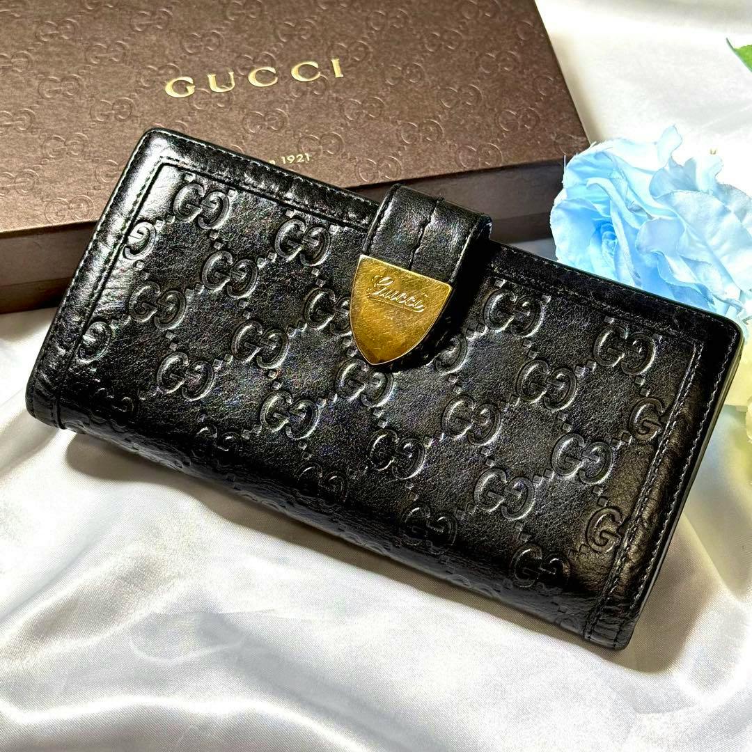 Black Gucci long flap wallet with embossed GG pattern and a gold Gucci clasp, photographed on fabric with a Gucci box in the background.