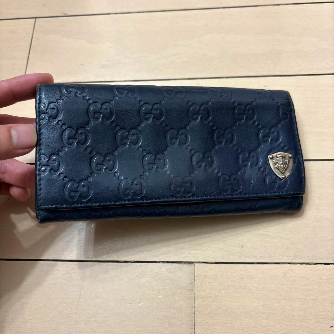 Closed navy GUCCI Guccissima embossed leather flap wallet with repeating GG pattern and a small metal shield emblem, held by a hand on a wooden floor.