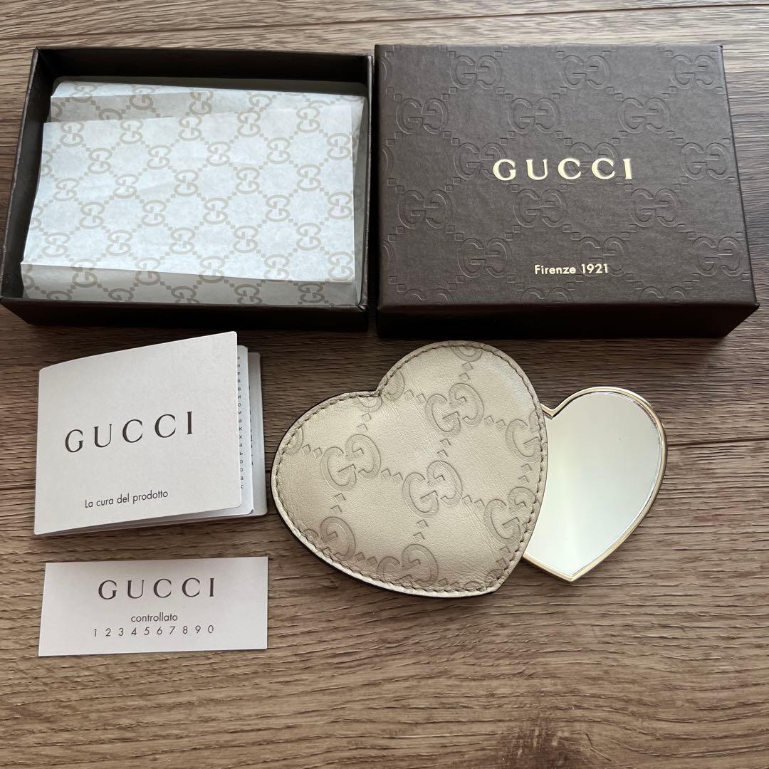 Heart-shaped Gucci compact mirror with GG monogram shown open beside its box and paperwork on a wooden surface.