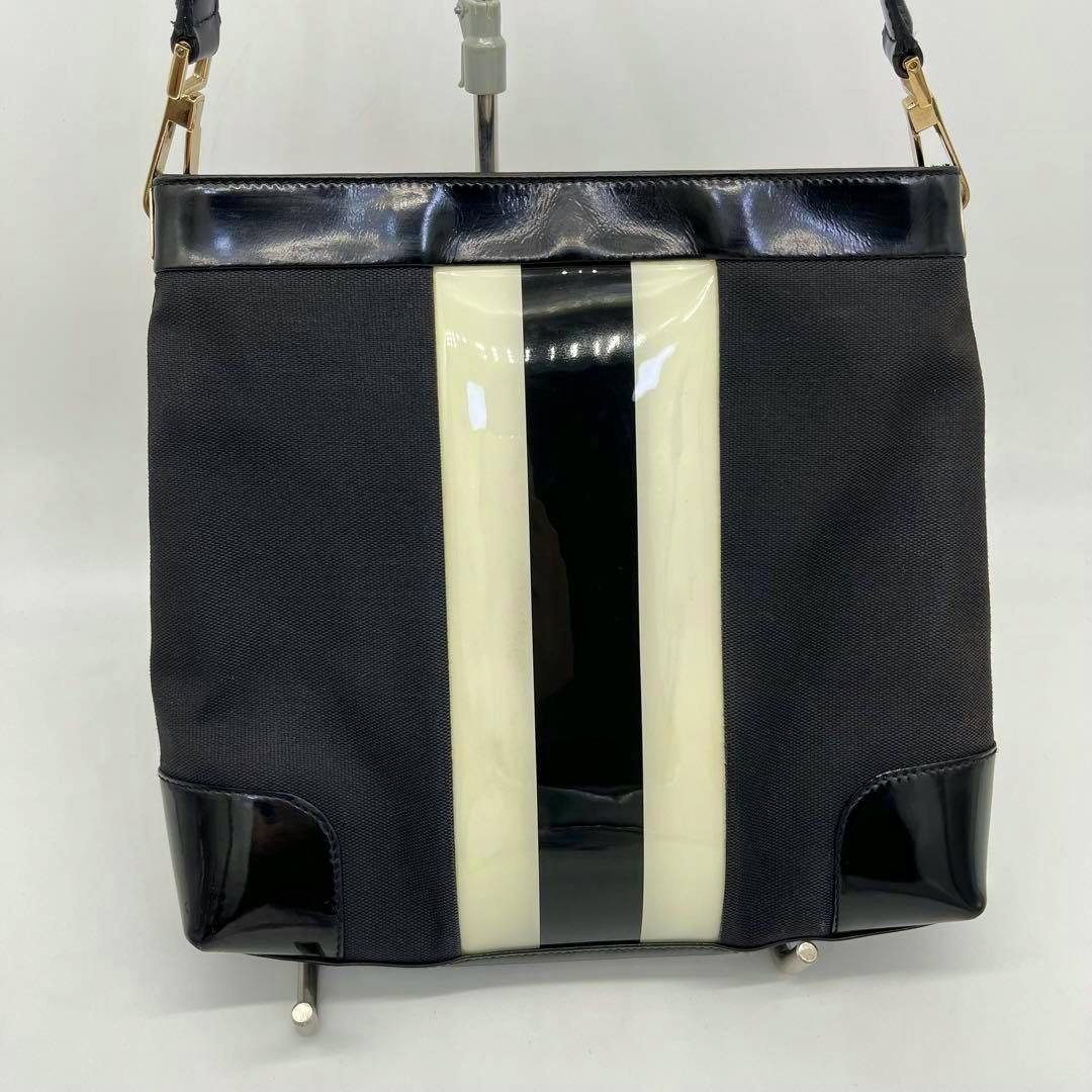 Front view of a black Gucci one-shoulder canvas bag with a central black-and-white enamel stripe and black patent leather trim.