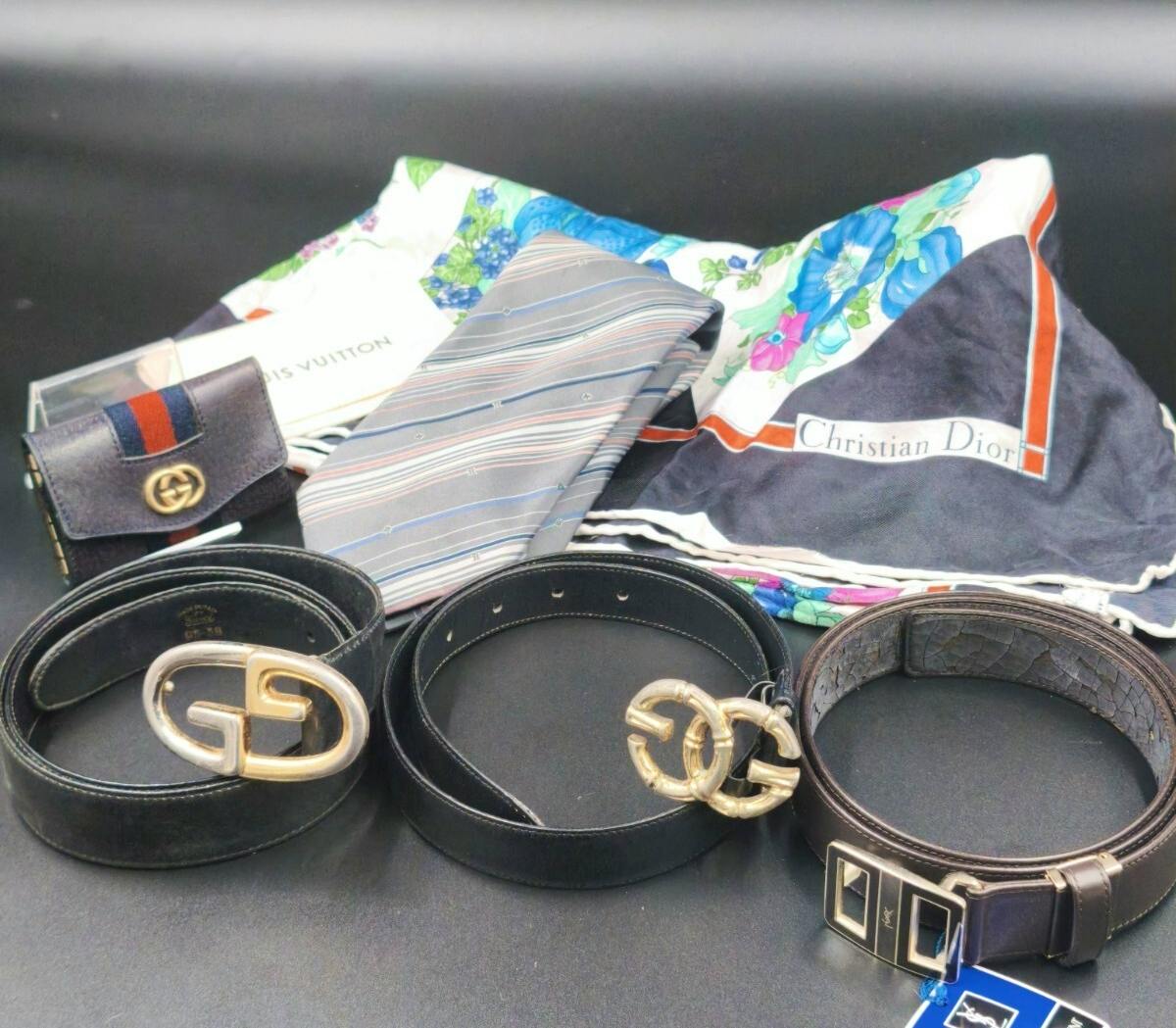 Several leather belts with metal buckles displayed alongside scarves, a tie, and a small key case on a dark background.