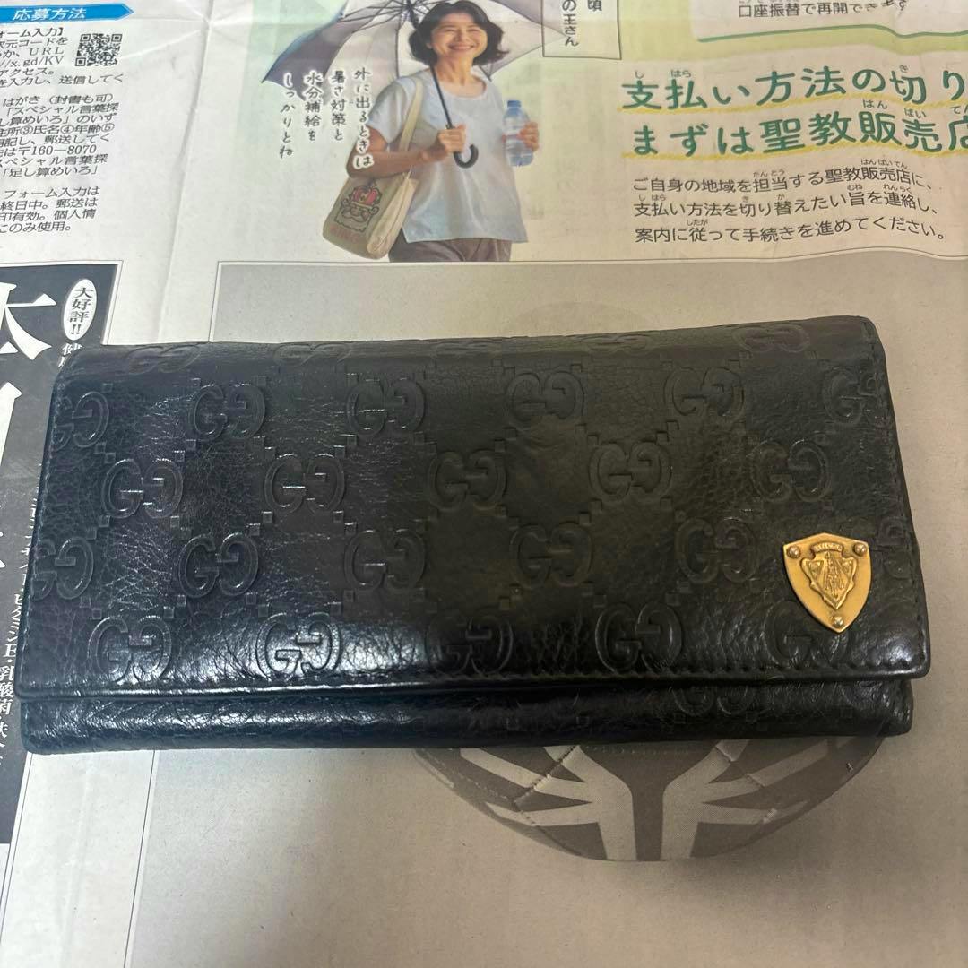 Black Gucci men's long wallet with embossed GG pattern and a gold shield emblem lying on newspaper.