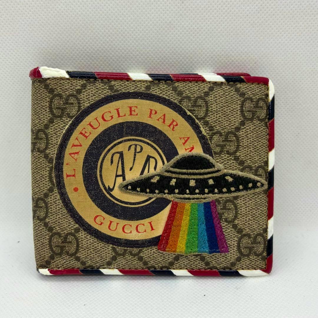 Front view of a closed brown Gucci two-fold wallet in GG monogram canvas with a UFO patch emitting a rainbow.