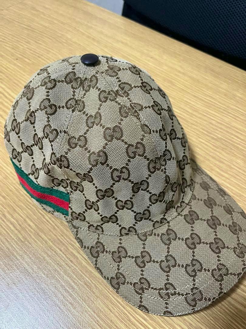Beige Gucci cap with all-over GG monogram pattern and a green-red side stripe on a wooden surface.