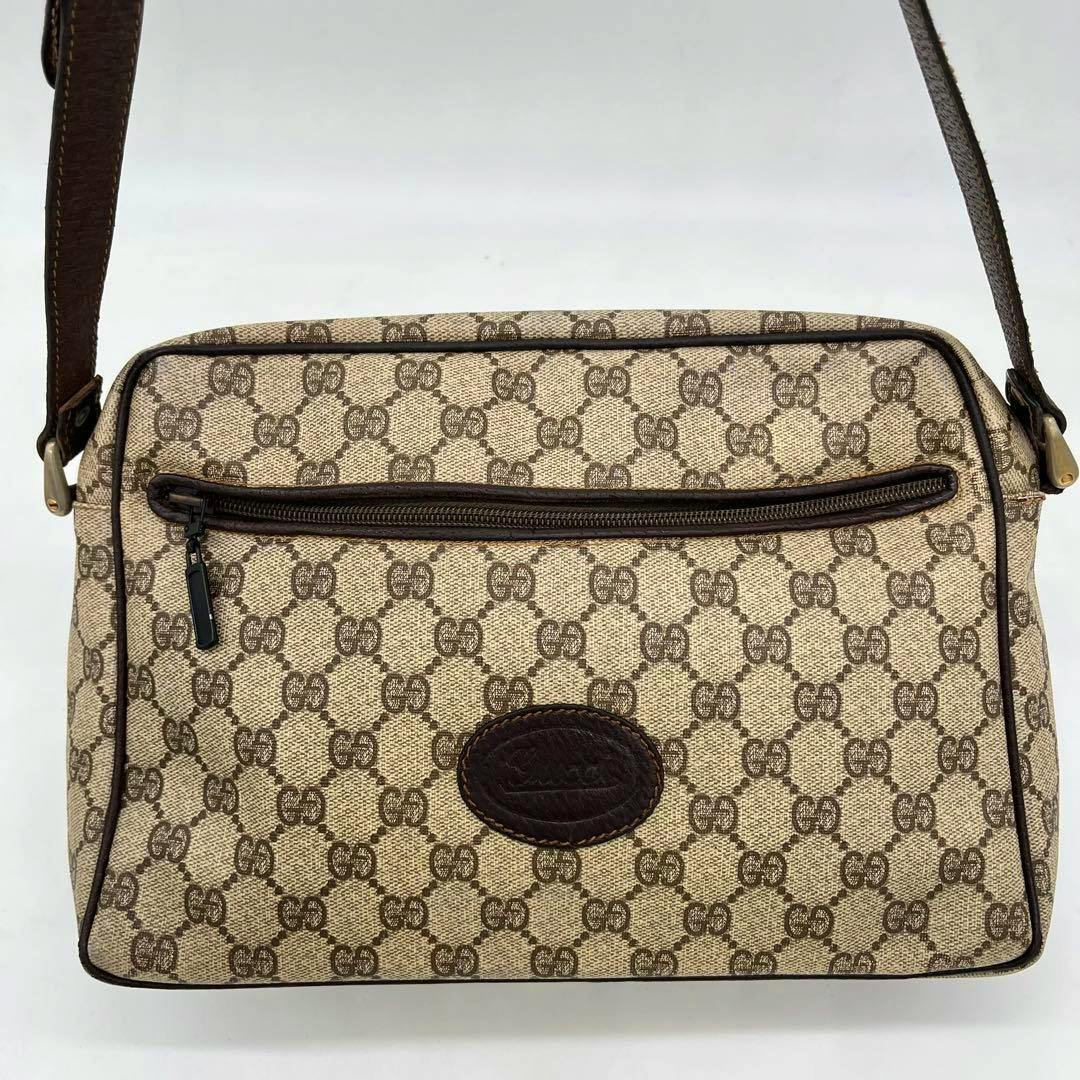 Front view of a GUCCI GG monogram canvas shoulder camera bag with a front zip pocket, leather oval logo patch, and brown leather strap.