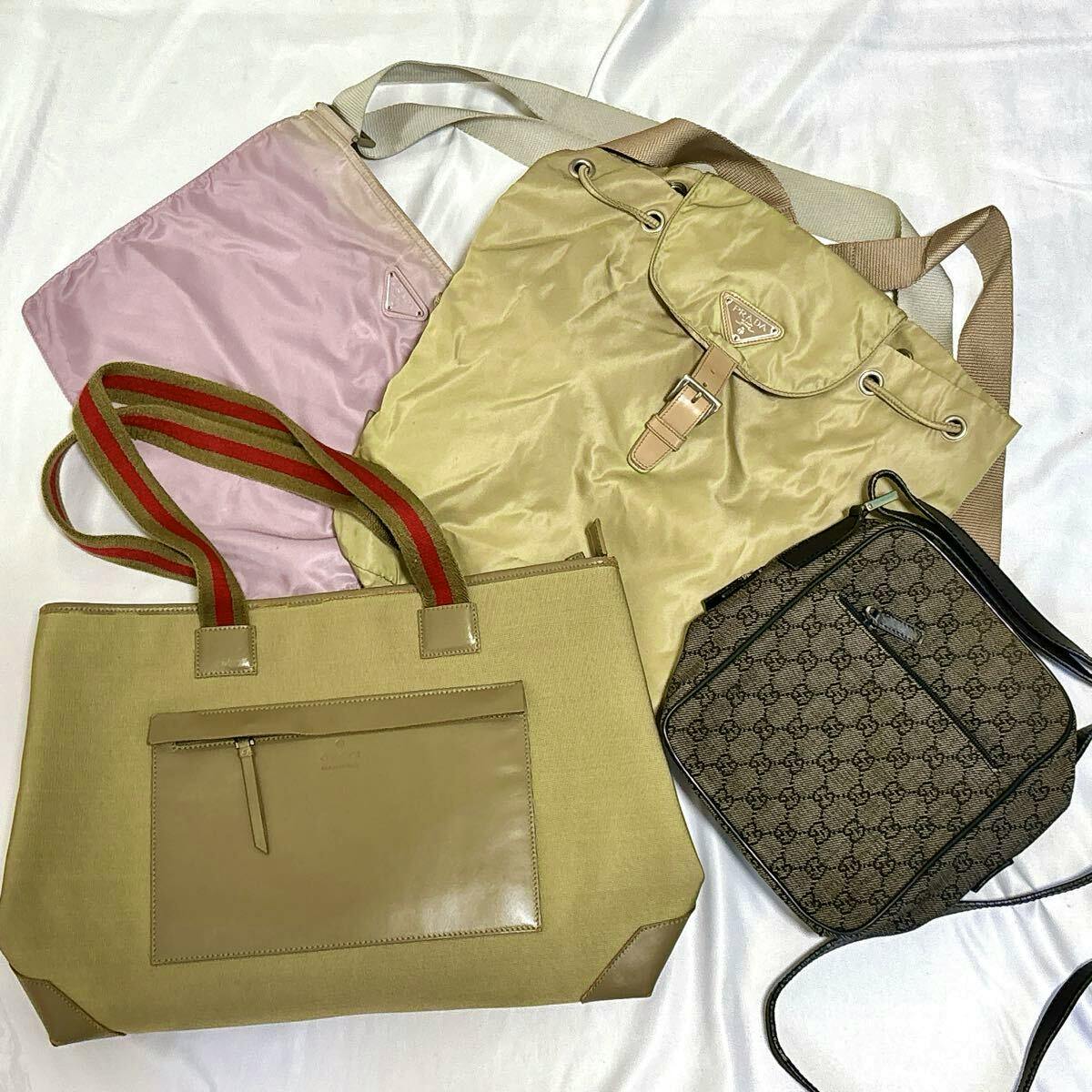 Four bags laid on a white surface: a beige tote, a beige Prada drawstring backpack, a pink pouch, and a brown Gucci monogram crossbody.