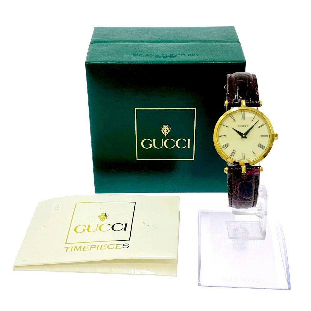 GUCCI ladies quartz wristwatch with white dial and gold-colored case displayed on a stand beside a green GUCCI box and booklet.