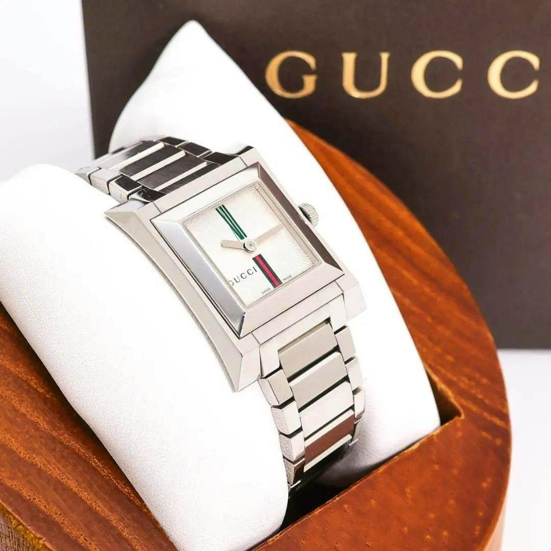 Silver square Gucci wristwatch with a metal bracelet and green-red stripe on the dial, displayed on a white cushion with a brown Gucci box partially visible in the background.