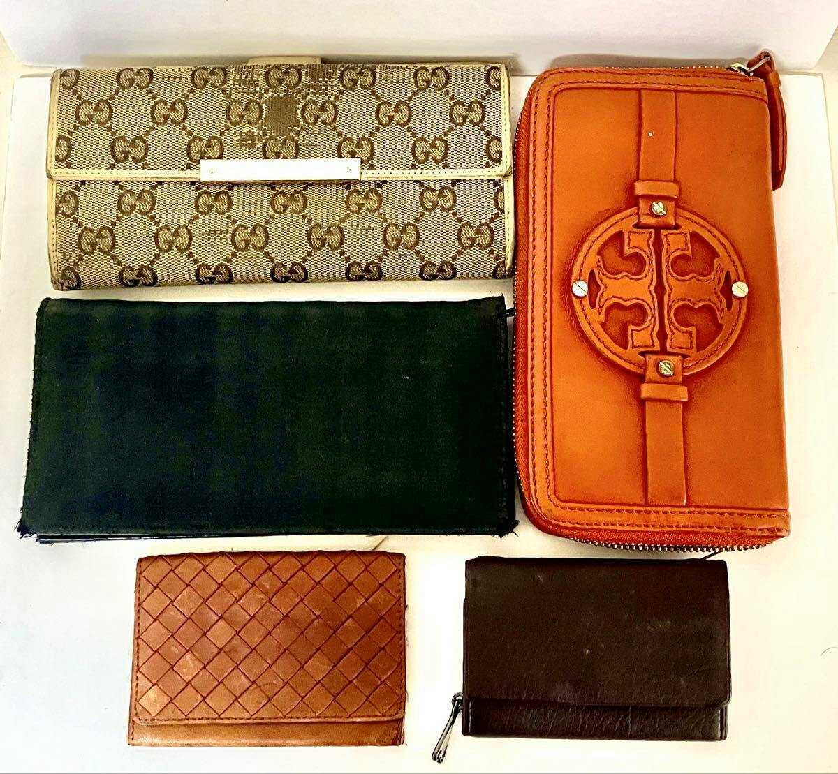 Five wallets and small leather goods arranged on a white background, including a beige Gucci monogram canvas wallet at top left and an orange zip-around wallet at top right.