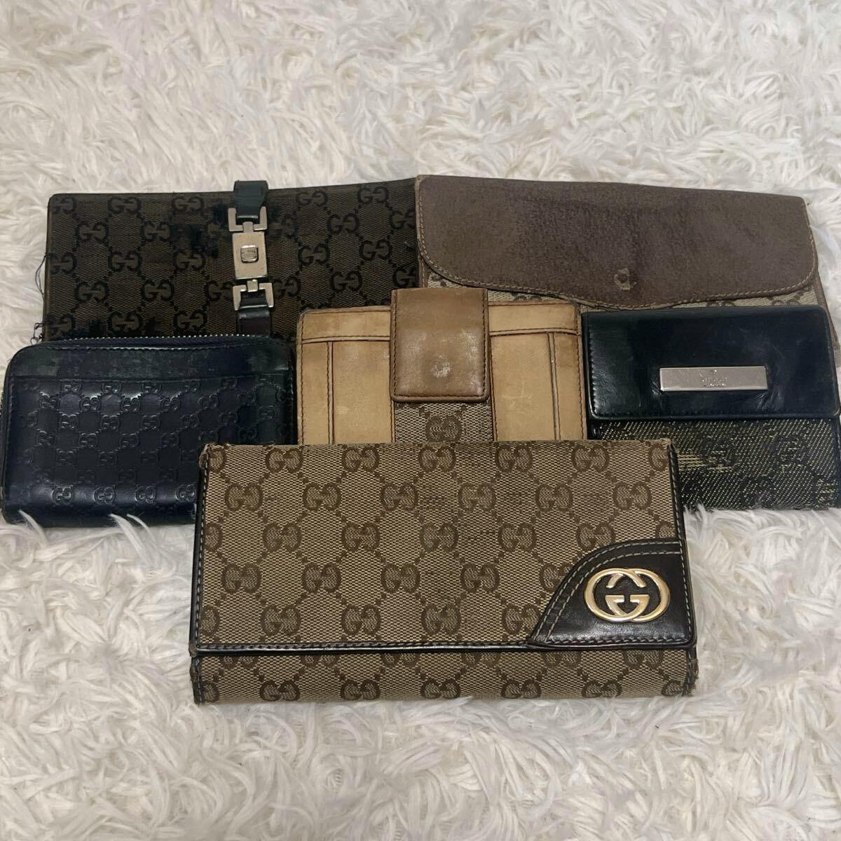 Six Gucci wallets in various styles and materials arranged on a white fuzzy surface, with a brown GG canvas long wallet prominent in front.