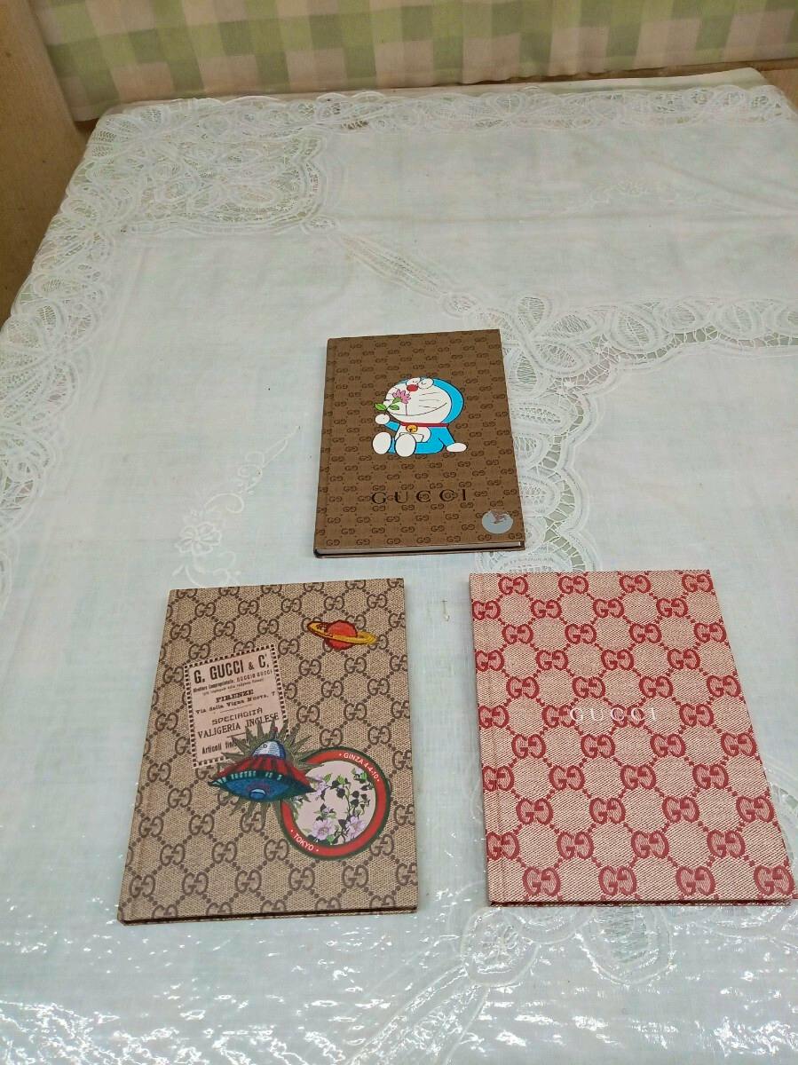 Three GUCCI-patterned notebooks, one showing a Doraemon illustration, arranged on a white lace tablecloth.