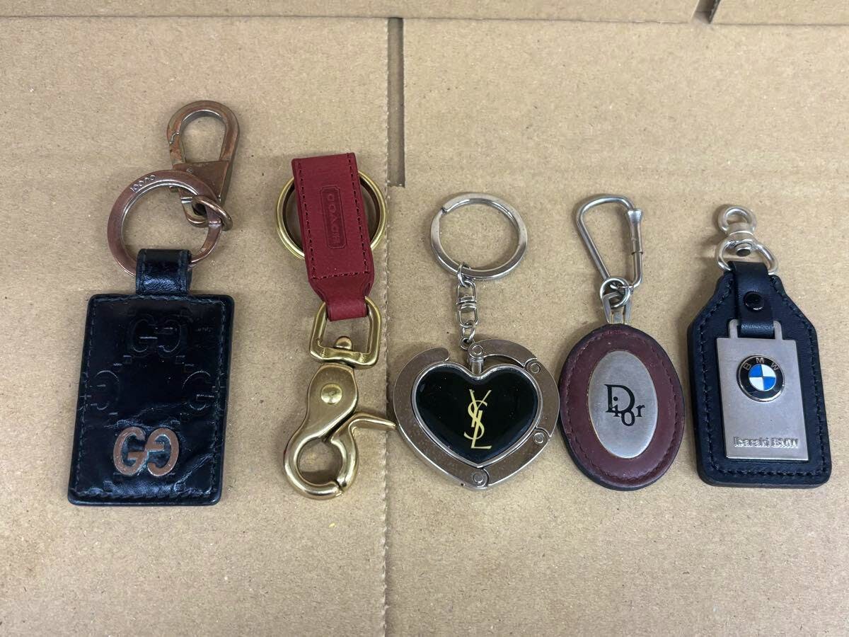 Five branded key holders and bag charms (GUCCI, COACH, YSL, Dior, BMW) laid side by side on a cardboard surface.