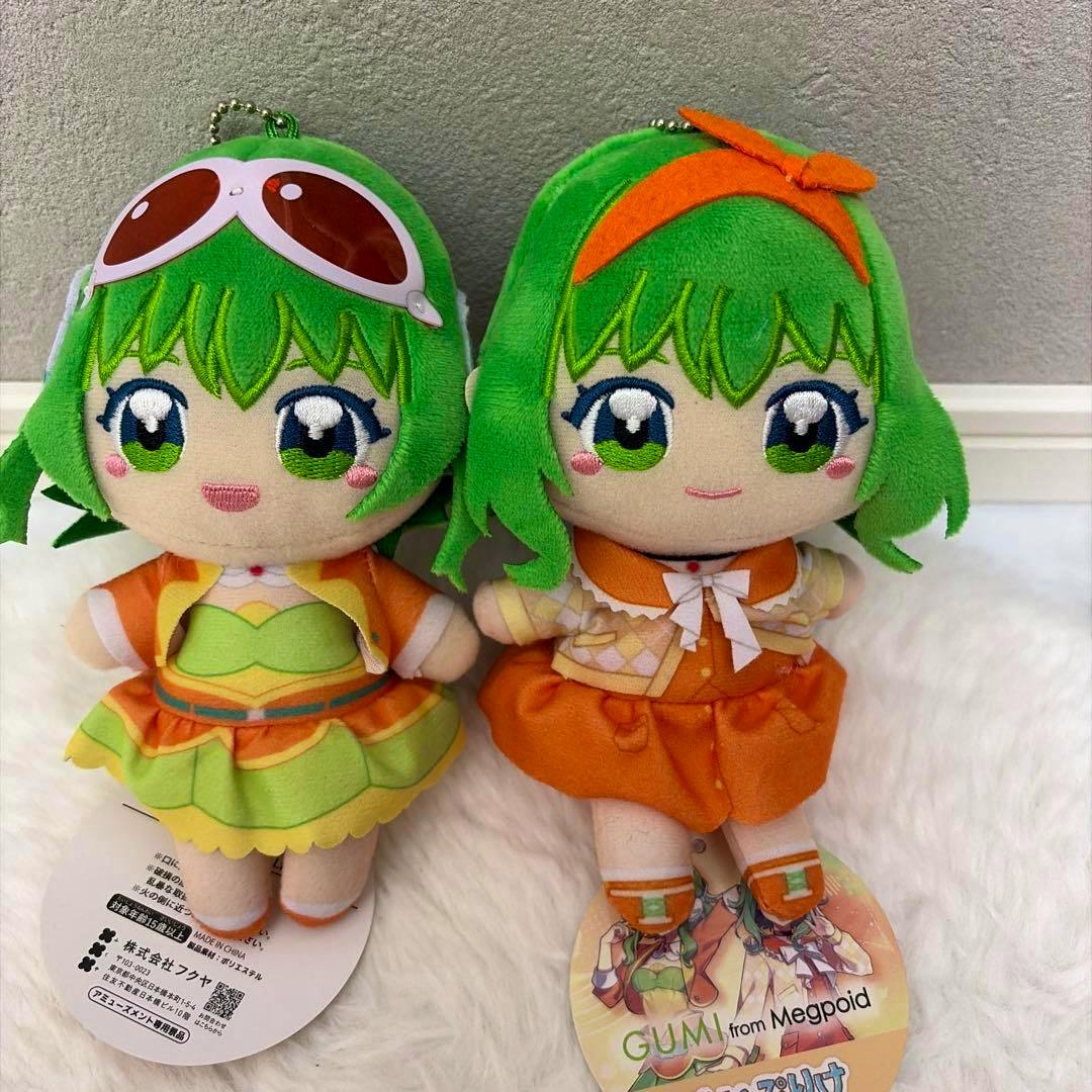 Two GUMI plush toys with green hair wearing orange outfits are shown sitting side by side on a white surface.
